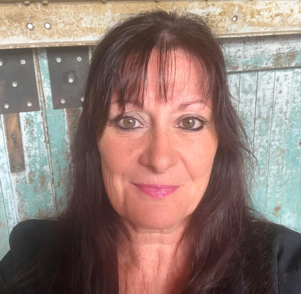 Portrait souriant d'une femme aux cheveux foncés et maquillage discret. Elle est devant un fond rustique de bois patiné et métal vieilli.