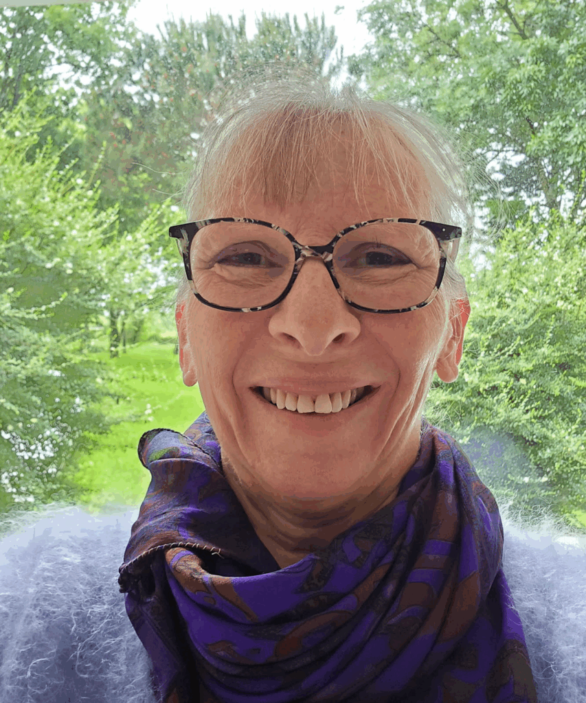 Femme âgée aux cheveux gris et lunettes, souriant joyeusement. Elle porte une écharpe violette et un pull bleu. Arbres verts en arrière-plan.