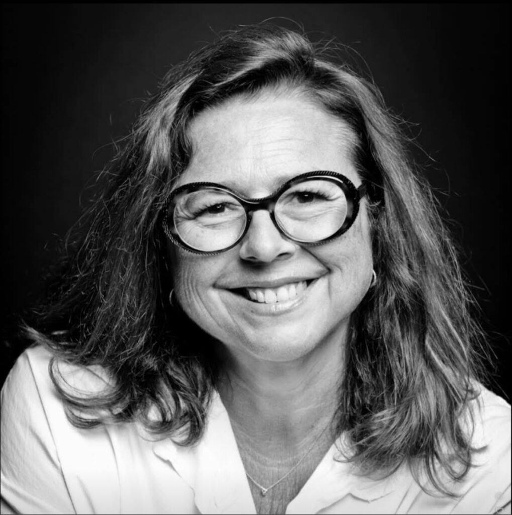 Portrait joyeux d'une femme aux lunettes rondes et cheveux ondulés, souriant avec optimisme en chemise blanche, noir et blanc.